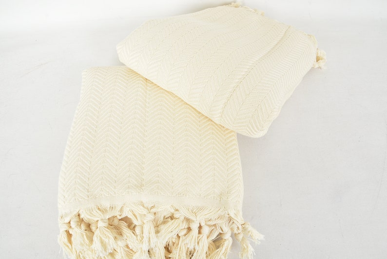 May include: Two cream-colored cotton towels with a herringbone pattern and fringe edges. The towels are folded and stacked on top of each other.
