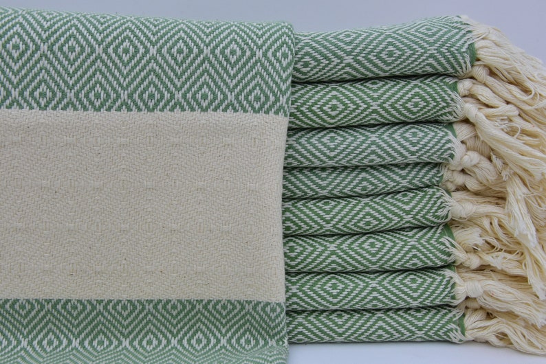 May include: A stack of green and white patterned Turkish towels with fringe. The towels are folded and stacked on a white surface.