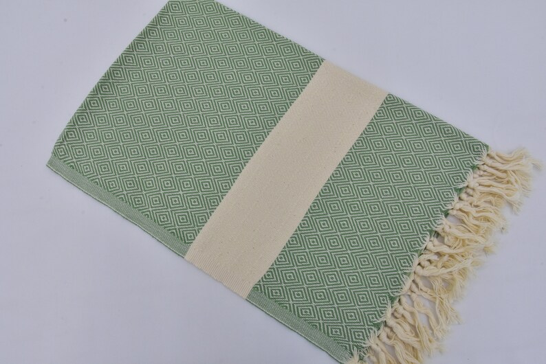May include: A green and white patterned Turkish towel with fringe. The towel has a diamond pattern and a white stripe down the center.