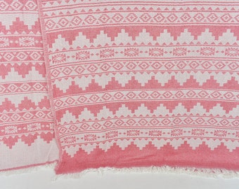 Puede incluir: Una toalla turca de color rosa y blanco con un borde de flecos. La toalla presenta un diseño geométrico con detalles blancos sobre un fondo rosa.