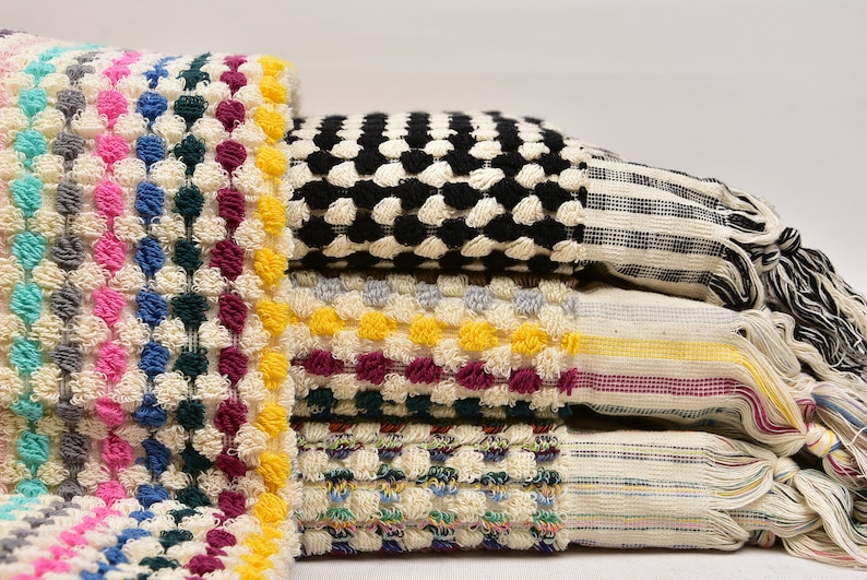 May include: A stack of colorful towels with a textured, puffy design. The towels are in various colors, including black, white, yellow, pink, blue, and green. The towels have fringe edges.