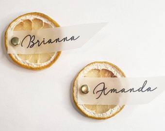 May include: Two dried lemon slices with gold tacks and white paper place cards with the names Brianna and Amanda written in black calligraphy.