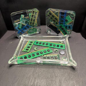 May include: Clear resin art pieces containing computer components. Two square blocks hold a mix of green and blue circuit boards. A rectangular tray displays green RAM sticks and small circular components, all encased in clear resin.