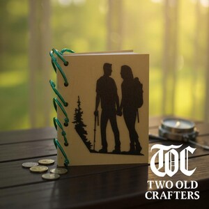 May include: A handmade journal with a hiking couple silhouette and a tree design. The journal is bound with green cord and features the text "TWO OLD CRAFTERS". The journal is on a wooden surface with coins and a compass.
