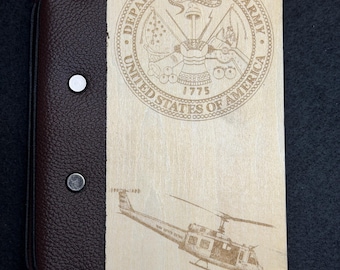 May include: A rectangular notebook with a light wood cover featuring an engraved Department of the Army seal and a helicopter. The spine is made of dark brown textured leather secured with four silver-toned metal rivets along the left edge.