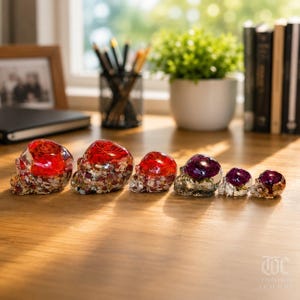 May include: A row of six skull-shaped resin ornaments of varying sizes, arranged from largest to smallest. The three larger skulls contain red floral elements and multi-colored stone chips, while the three smaller skulls feature purple floral accents.
