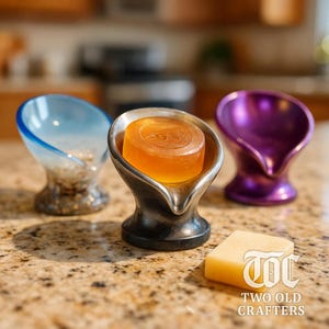 May include: Three soap dishes in blue, silver, and purple, each holding a bar of soap. The silver dish holds an orange soap bar. A separate bar of soap with the text "TWO OLD CRAFTERS" is also present.