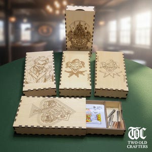 May include: Wooden playing card boxes with engraved designs. The designs include a Joker, a Spade, and playing card suits. One box is open, revealing playing cards and metal accessories. The logo "TWO OLD CRAFTERS" is visible.