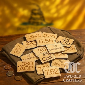 May include: A collection of small, rectangular wooden plaques with various numerical and textual markings, including "357", "5.56", and "45 ACP". The plaques are arranged on a folded brown fabric, with a few coins visible. The background features a yellow flag.