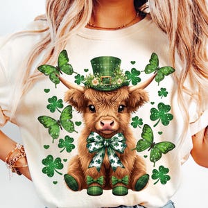 May include: A cream-colored t-shirt featuring a brown Highland cow wearing a green leprechaun hat adorned with flowers. The cow has a green and white bow tie and plaid shoes. Green butterflies and shamrocks surround the cow.