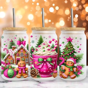Könnte beinhalten: Drei Glasbecher mit Bambusdeckeln und Metallstrohhalmen. Jeder Becher hat ein festliches Weihnachtsdesign mit Lebkuchenhäusern, Weihnachtsbäumen und Lebkuchenmännern. Die Designs beinhalten rosa, grüne und weiße Farben, ideal für Weihnachtsgetränke.