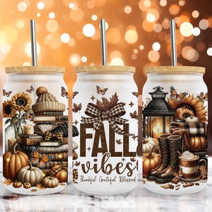 Könnte beinhalten: Drei Glasbecher mit Holzdeckeln und Metallstrohhalmen. Jeder Becher hat ein Herbstmotiv mit Kürbissen, Sonnenblumen und dem Text "Fall Vibes". Die Designs umfassen Schmetterlinge, Bücher und eine Laterne.