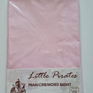 May include: A package of two flat percale sheets in a light pink color, designed for a pram, crib, or Moses basket. The packaging features the "Little Pirates" logo, a map, and a drawing of a ship.