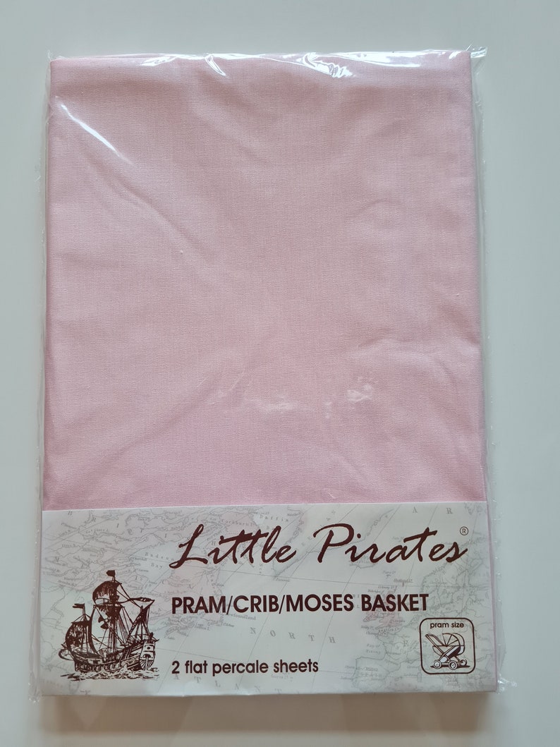 May include: A package of two flat percale sheets in a light pink color, designed for a pram, crib, or Moses basket. The packaging features the "Little Pirates" logo, a map, and a drawing of a ship.