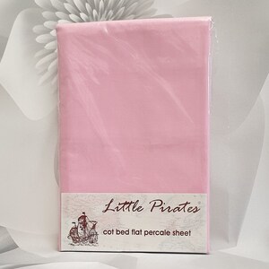May include: A flat pink cot bed sheet, packaged in clear plastic. The label reads "Little Pirates" and "cot bed flat percale sheet". The background features a blurred floral design in shades of grey and white.