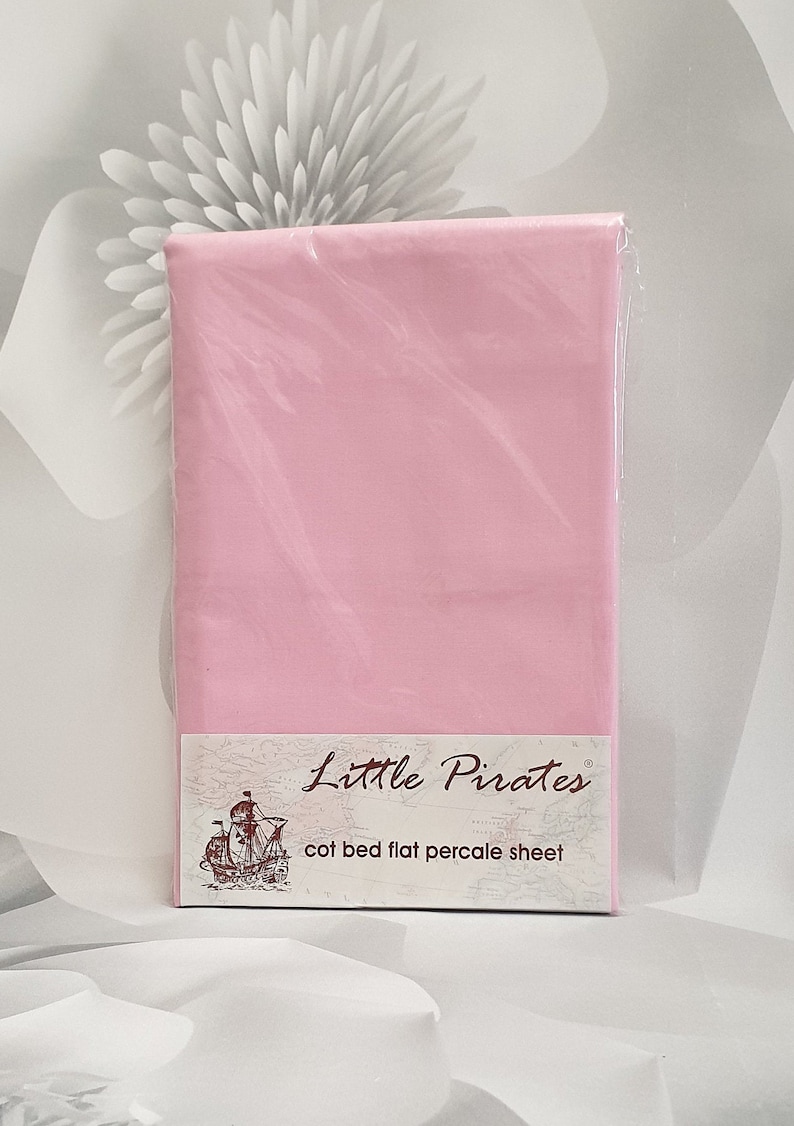 May include: A flat pink cot bed sheet, packaged in clear plastic. The label reads "Little Pirates" and "cot bed flat percale sheet". The background features a blurred floral design in shades of grey and white.