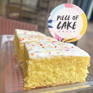 Puede incluir: Primer plano de una porción de pastel amarillo con glaseado blanco y chispas de colores. El pastel está en un recipiente de plástico transparente. La imagen incluye un logotipo circular que dice "PIECE OF CAKE OMAGH".