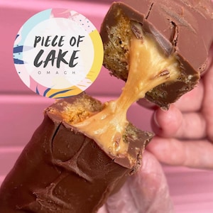 Puede incluir: Una barra de chocolate partida por la mitad, revelando un relleno de caramelo y un interior similar a un pastel. La barra de caramelo se sostiene sobre un fondo rosa. Un logotipo circular con el texto "PIECE OF CAKE OMAGH" está en la esquina superior izquierda.