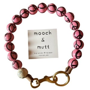 Op de afbeelding: Roze kralen hondenhalsband met vredesymbolen geschilderd op elke kraal. De halsband heeft een gouden metalen sluiting en een klein label met de tekst "mooch & mutt hip scrubs & hip pups venice, ca."