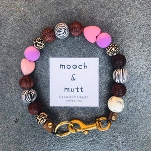 May include: A brown leather dog collar with a gold clasp. The collar is decorated with brown wooden beads, pink heart-shaped beads, and beads with zebra and leopard print patterns. The collar has a tag that reads "mooch & mutt big snacks & big pups vermont, vt."