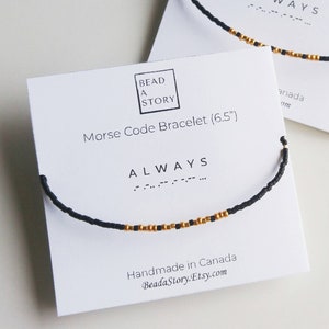 May include: Two white cards with black text and a black and gold beaded bracelet on each card. The text on the cards reads "Bead a Story" and "Morse Code Bracelet" with the size of the bracelet in inches. The word "ALWAYS" is written in Morse code on each card.