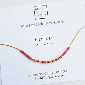 May include: A gold chain necklace with pink beads spelling out the name "ÉMILIE" in Morse code. The necklace is on a white card with the text "Bead a Story Morse Code Necklace".