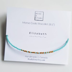 May include: A turquoise blue beaded bracelet with gold beads spelling out the name "Elizabeth" in Morse code. The bracelet is 8.5 inches long and is handmade in Canada. The card reads "Bead a Story" and "Morse Code Bracelet (8.5")".