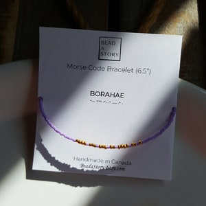 May include: A purple and gold beaded bracelet with the word "BORAHAE" written in Morse code on a white card. The card also says "Morse Code Bracelet (6.5")" and "Handmade in Canada BeadaStory.Etsy.com".