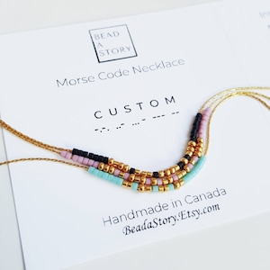 Peut inclure: Un collier en chaîne dorée avec des perles noires, roses et turquoise. Le collier est disposé en forme de courbe et épelle le mot "CUSTOM" en code Morse. Le texte "BEAD A STORY" et "Morse Code Necklace" est imprimé sur une carte blanche.