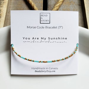 Peut inclure: Un bracelet en perles turquoise et dorées avec le message "You Are My Sunshine" en code Morse sur une carte blanche. La carte indique également "Morse Code Bracelet (7")" et "Handmade in Canada".