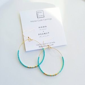 Peut inclure: Une paire de boucles d'oreilles créoles turquoise et or avec les mots "MAMA + PEANUT" en code Morse sur une carte blanche. La carte indique également "Bead a Story" et "Fabriqué au Canada".