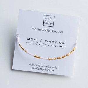 May include: White and gold beaded bracelet with the words "MOM / WARRIOR" in Morse code. The bracelet is on a white card with the text "BEAD A STORY Morse Code Bracelet Handmade in Canada BeadaStory.Etsy.com"