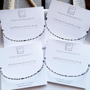 Peut inclure: Quatre cartes blanches avec des bracelets bleus et argentés. Chaque carte porte le texte "Bead a Story" et "Bracelet en code Morse" avec la taille du bracelet en pouces. Le texte "California Girls Trip" est également présent sur chaque carte.