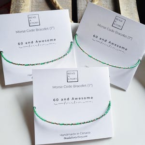 Peut inclure: Trois cartes blanches avec des bracelets en perles vertes et dorées. Chaque carte porte le texte "Bead a Story" et "Bracelet en code Morse (7')" et "60 et Awesome" avec le code Morse de la phrase. La carte du bas porte également le texte "Fabriqué au Canada" et "Beadastory.Etsy.com".