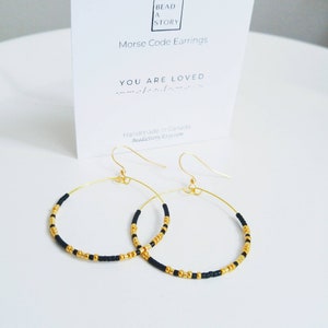May include: Gold hoop earrings with black beads. The earrings are handmade in Canada and feature the message "You are loved" in Morse code.