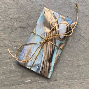 Pode incluir: Um pequeno pacote embrulhado em papel estampado azul e marrom com detalhes dourados. O pacote &eacute; amarrado com um barbante natural.
