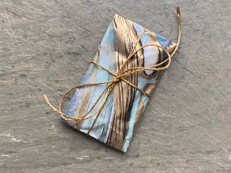 Pode incluir: Um pequeno pacote embrulhado em papel estampado azul e marrom com detalhes dourados. O pacote &eacute; amarrado com um barbante natural.