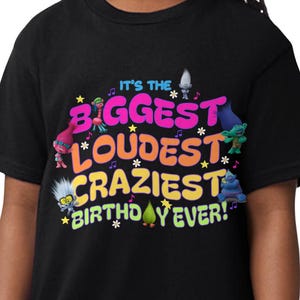 May include: A black t-shirt featuring colorful text and graphics. The text reads "It's the Biggest, Loudest, Craziest Birthday Ever!" with images of cartoon characters. The shirt is worn by a person with braided hair.