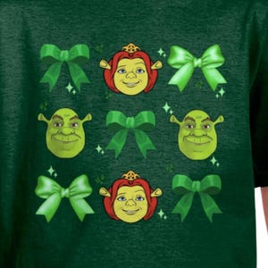 May include: Green t-shirt featuring a design with green bows, and cartoon faces of Shrek and Princess Fiona. The design includes sparkling accents. The shirt is made of a dark green fabric.