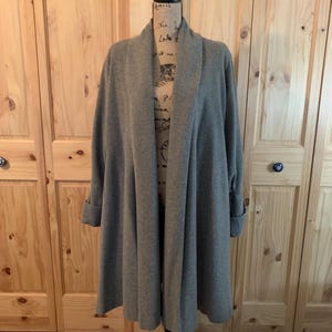 May include: A long, open-front gray cardigan displayed on a mannequin. The cardigan has a shawl collar, long sleeves with folded cuffs, and a flowing, knee-length design. The mannequin is on a black stand, set against a wooden background.