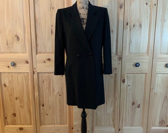 May include: A black double-breasted coat with a long, straight silhouette. The coat is on a black mannequin and has a simple, classic design.