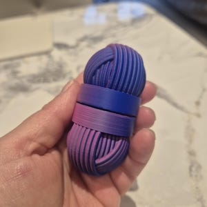 Puede incluir: Una madeja de hilo impresa en 3D en tonos azules y rosas, sostenida en una mano. La madeja es cilíndrica con estrías horizontales y está dividida por una banda de los mismos colores. Es probable que sea un objeto decorativo o un suministro de artesanía.