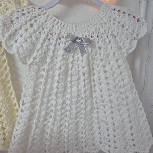 May include: White crocheted baby dress with a silver bow and pink trim. The dress has a scalloped hem and short sleeves.