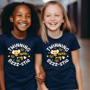 Può includere: Due bambini indossano magliette abbinate blu navy con un'immagine di un'ape gialla e il testo "TWINNING with my Bzzz-stie".