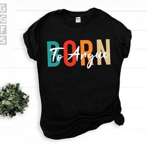 Pode incluir: Camiseta preta com a frase "Born To Argue" em letras de bloco turquesa, vermelho, laranja e bege. A palavra "To" está em escrita branca. Uma pequena planta está no canto inferior esquerdo.