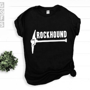 Puede incluir: Camiseta negra con la palabra "ROCKHOUND" en blanco y un gráfico de pico blanco. La camiseta de cuello redondo está hecha de tela oscura. El diseño probablemente está dirigido a personas interesadas en geología o coleccionismo de rocas.