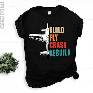 May include: Black t-shirt with a white silhouette of a plane and the text "BUILD FLY CRASH REBUILD" in colorful retro lettering.