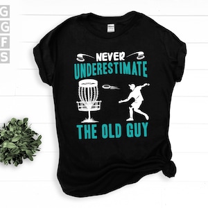 May include: Black t-shirt with a white graphic of a disc golf player throwing a disc at a basket. The text "Never underestimate the old guy" is printed in teal.