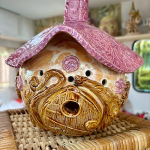 Könnte beinhalten: Ein skurriles Vogelhaus aus Keramik mit rosa Dach und Schornstein und einem beigefarbenen Körper. Das Vogelhaus hat eine detaillierte Tür, florale Akzente und dekorative Muster. Es steht auf einem geflochtenen Korb.