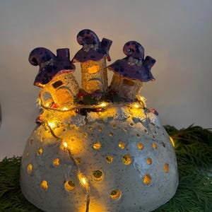 Könnte beinhalten: Ein Keramik-Feenhaus mit drei Häusern mit violetten Dächern und weißen Akzenten und einer Lichterkette um die Basis. Die Häuser stehen auf einer weißen Keramikbasis mit Löchern.
